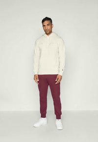 Hoddy beige chiaro con logo frontale e cappuccio regolabile, abbinato a pantaloni della tuta bordeaux e sneakers bianche, con polsini a coste.