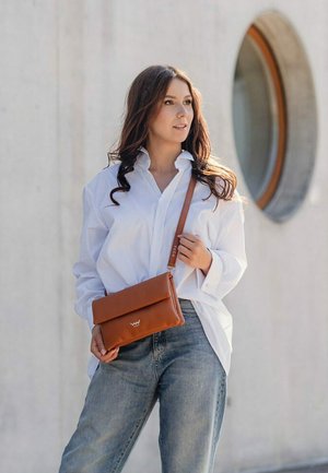 Hnědá kožená crossbody taška s hladkým povrchem a klopou. Model má na sobě bílou oversized košili a světlemodré džíny.