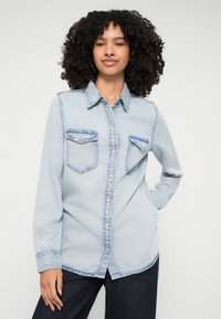 Light blue denim shirt featuring a pointed collar, front button closure, and two chest pockets with contrasting stitching. Long sleeves with buttoned cuffs.