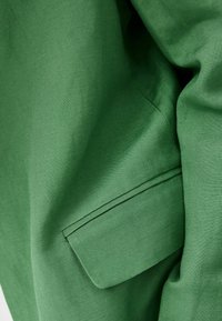 Zbliżenie na zielony materiał, ukazujące kieszeń z klapą, podkreślające fakturę i detale przeszycia materiału.