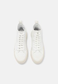 Paire de baskets montantes en toile blanche avec des lacets blancs et des semelles en caoutchouc beige, vues de dessus sur un fond blanc.