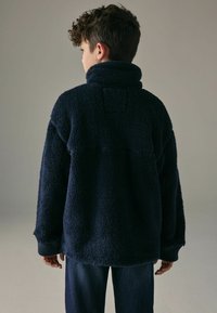 Pullover in pile blu navy con colletto alto, superficie testurizzata, vestibilità oversize e polsini a costine. La vista posteriore mostra un design senza cuciture.