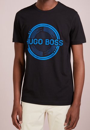 Czarny t-shirt z krótkim rękawem z niebieskim okrągłym wzorem i tekstem "HUGO BOSS", noszony przez osobę w beżowych spodniach.