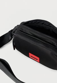 Melna auduma soma pāri plecam ar fakturētu virsmu, divām kabatām ar rāvējslēdzējiem un sarkanu logotipa uzlīmi. Komplektā ir regulējams plecu siksna.