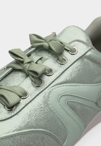 Gros plan sur une basket vert clair à éclat métallique, avec de larges lacets texturés noués en nœuds, des œillets argentés et des détails de couture courbés sur le côté.