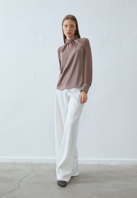 Brown blouse with a keyhole neckline and long sleeves; paired with white wide-leg trousers and dark gray shoes on a light background.