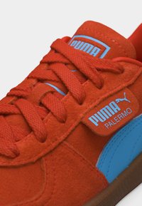 Sneaker Puma Palermo in suede rosso con logo blu, striscia laterale blu e lacci rossi abbinati su suola marrone.