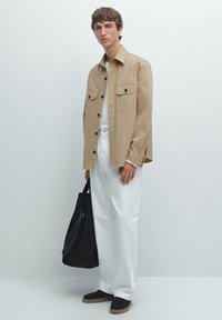 Camicia beige con bottoni, due tasche sul petto, t-shirt bianca e pantaloni bianchi larghi. Scarpe slip-on in suede nero e una borsa a tote scura.