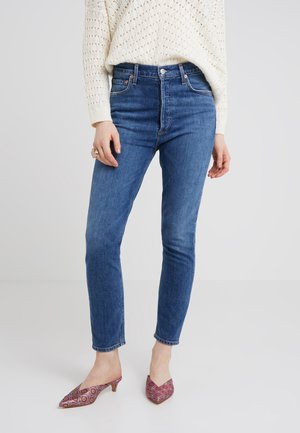 Femme portant un jean skinny bleu taille mi-haute, un pull en maille crème et des mules à bout pointu à talons bas et motifs, posant sur un fond uni.