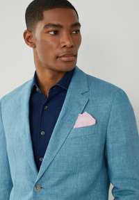 Blazer in lino azzurro chiaro con rever a punta, abbinato a una camicia color blu scuro con colletto e un fazzoletto da taschino rosa con motivo a pois bianchi.
