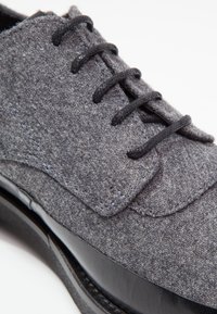 Šedá plstěná bota s černými tkaničkami, s hladkou černou koženou podrážkou a texturovaným svrškem. Kulatá špička a šitý detail.