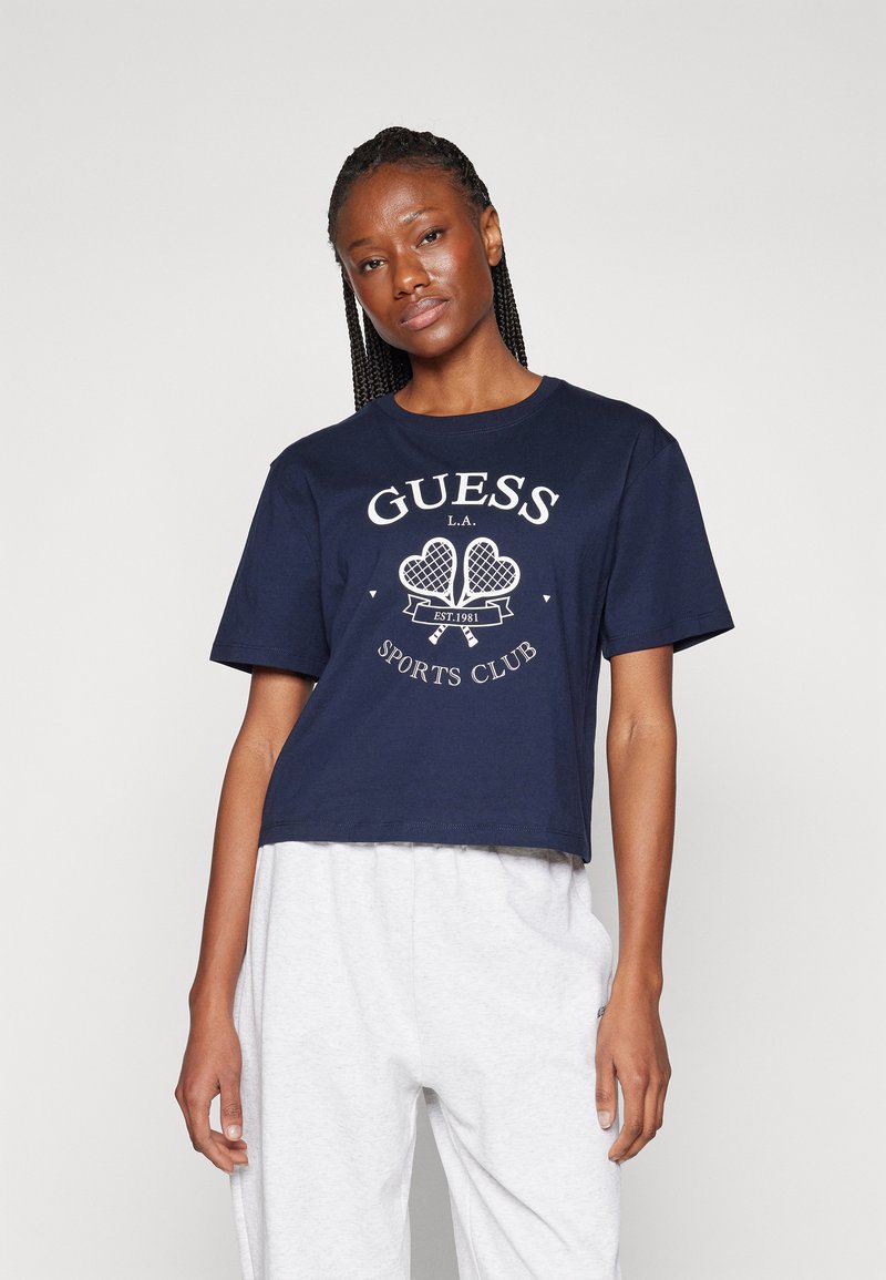 Lähinä navy-sininen puuvilla cropped t-paita, jossa on valkoinen graafinen printti, jossa lukee "GUESS L.A. SPORTS CLUB" ja tennismailan malli. Lyhyet hihat.