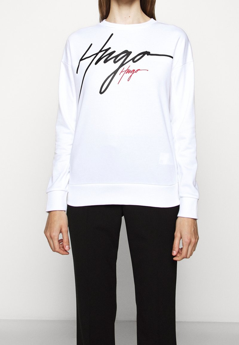 Personne portant un sweat-shirt blanc à manches longues avec un grand logo "Hugo" en lettres noires et rouges sur le devant, associé à un pantalon noir.