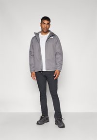 Grå vandtæt jakke med hætte, elastiske manchetter. Båret over en hvid t-shirt, parret med mørke slim-fit bukser og grå vandresko.