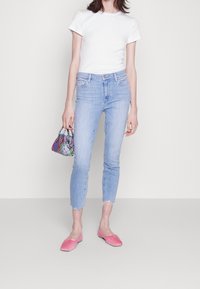 Žena v bílém přiléhavém tričku, světle modrých zkrácených džínech s roztrhanými lemy, růžových balerínách, držící malou kabelku s kostkovaným vzorem v různých barvách.