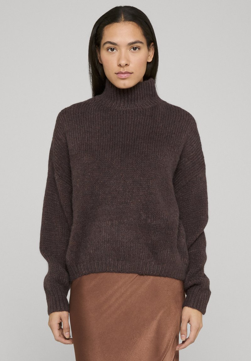 Oversized donkerbruin geribbelde gebreide trui met een hoge hals en lange mouwen, gecombineerd met een zijdezachte bruine rok. Zachte, gestructureerde stof.