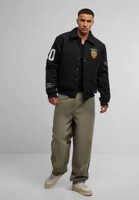 Black wool varsity jacket with logo details, large number '10' on the sleeve, paired with loose olive green pants and beige sneakers.