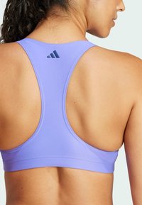 Ljubičasti sportski grudnjak s dizajnom racerback, izrađen od glatkog materijala. Karakterizira ga Adidas logo u tamnoplavoj boji na gornjem dijelu leđa.