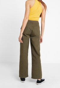 Pantaloni a palazzo verde oliva con vita alta, realizzati in tessuto resistente, abbinati a una canotta gialla a costine e sneakers nere.