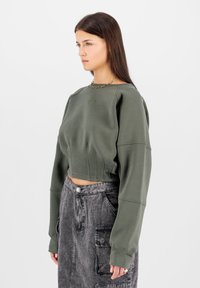 Rövidített, olívazöld pulóver bordás részletekkel és oversized ujjal, magas derékú, fakó fekete szoknyával, amely zsebekkel rendelkezik.