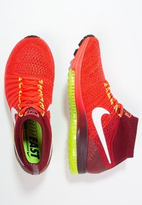 Červené pletené sportovní boty s vysokým designem, bílým logem Swoosh, žlutými tkaničkami a viditelnou vzduchovou odpruženou podrážkou.