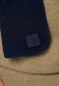 Mørkeblå strikket tørklæde med struktureret mønster, prydet med et rektangulært label i mørkeblått med prægede logo, lagt mod en beige sweater.