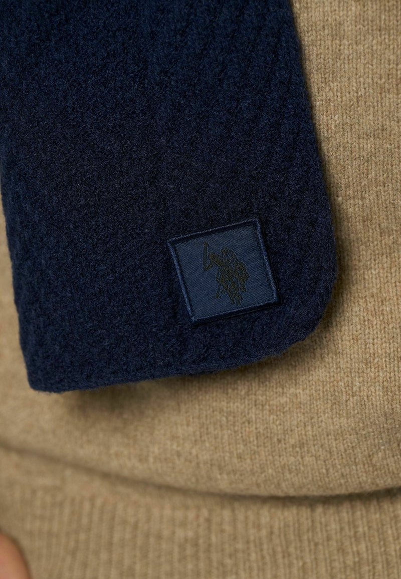 Mørkeblå strikket tørklæde med struktureret mønster, prydet med et rektangulært label i mørkeblått med prægede logo, lagt mod en beige sweater.