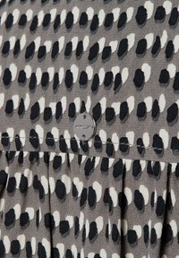 Grijze stof met een abstract zwart-wit stippenpatroon, voorzien van een klein, rond zilveren label met de merknaam "Marc Cain" erop genaaid.