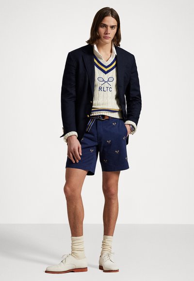 Jūras kruzu stila žakete, krēmveida adīta džemperis ar zilu akcentu, izšūti tenisa motīvi uz jūras ziliem šortiem, krēmveida zeķes un bēšas apavus.