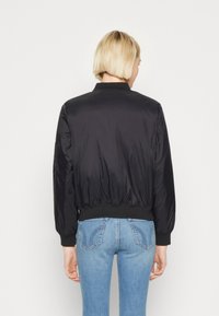 Blouson aviateur noir à texture lisse et brillante, avec des poignets et un ourlet côtelés. Porté avec un jean bleu clair ajusté, mettant en valeur une silhouette ajustée.