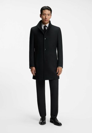 Mann, der einen schwarzen formellen Mantel über einem weißen Hemd und einer schwarzen Krawatte trägt, schwarze Hose und schwarze elegante Schuhe, stehend vor einem weißen Hintergrund.