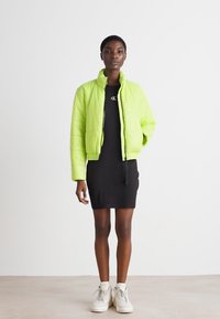 Veste courte matelassée verte néon avec une finition brillante, fermeture éclair et poignets élastiques, portée par-dessus une robe noire et des baskets blanches.