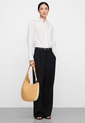 Woman standing wearing a white button-up shirt, black wide-leg pants, black belt, holding a large tan leather handbag with short handles.
