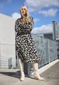 Vestido de leopardo en blanco y negro con mangas largas, de longitud hasta la rodilla, combinado con botas de punta beige hasta la rodilla. Entorno urbano al aire libre.