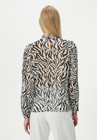 Durchsichtige Bluse mit einem schwarz-weißen Zebra-Muster, ausgestattet mit einem Schlüsselaugenverschluss am Rücken und langen Ärmeln mit gerafften Manschetten.