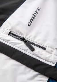 Giacca bianca e nera con tasca con zip, logo "ombre" in nero e una finitura liscia e opaca. Cuciture a contrasto visibili sui bordi.