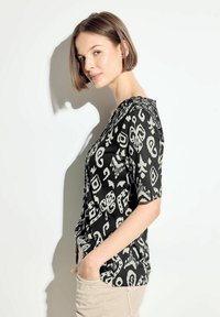 Camisa de patrón en blanco y negro con mangas cortas, que presenta un cuello redondo y un corte holgado. La tela parece ligera y texturizada.