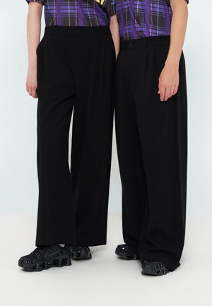 Deux personnes portant un pantalon large noir, des baskets épaisses noires et des chemises à carreaux violettes se tiennent côte à côte sur un fond clair.