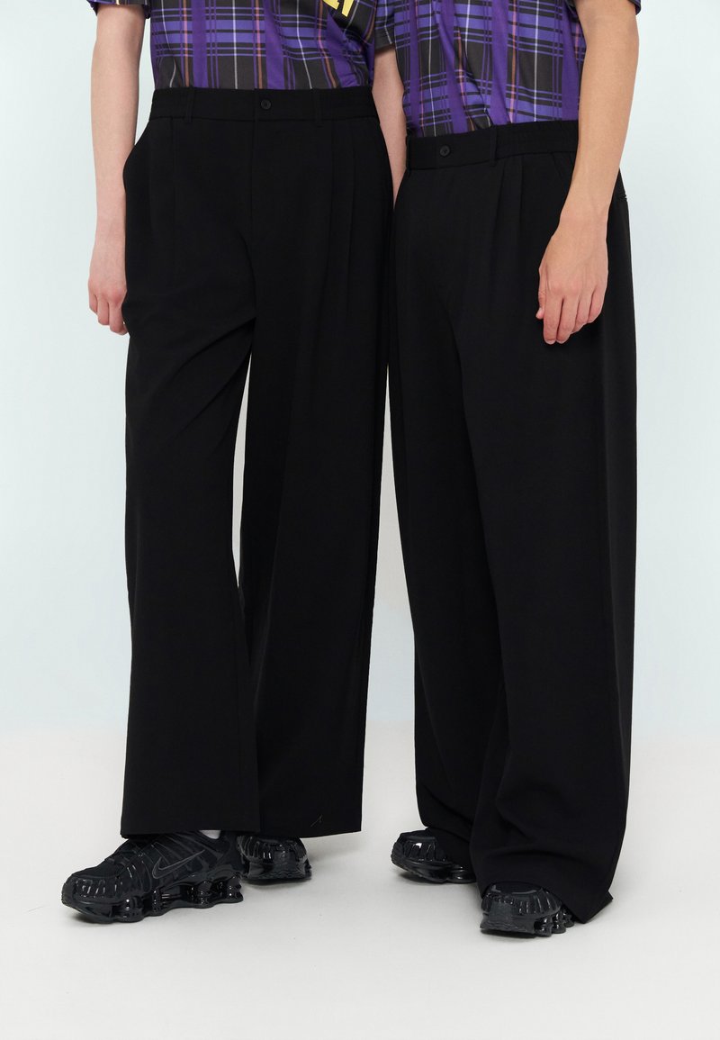 Deux personnes portant un pantalon large noir, des baskets épaisses noires et des chemises à carreaux violettes se tiennent côte à côte sur un fond clair.