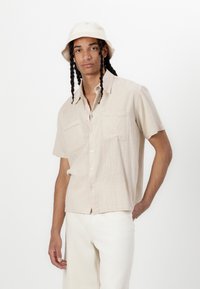 Camisa de manga corta de un beige claro con dos bolsillos en el pecho y un cierre de botones. Llevada con pantalones de color claro y un sombrero de cubo a juego.