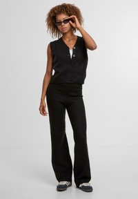 Černá pletená vesta s předním zapínáním na knoflíky, v kombinaci s volnými kalhotami ve stejném stylu. Modelka má na sobě tmavé sluneční brýle a tenisky, čímž předvádí minimalistický styl.