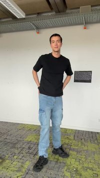 Un jeune homme se tient à l'intérieur contre un mur blanc avec du texte, portant un t-shirt noir, un pantalon cargo bleu clair et des baskets noires.