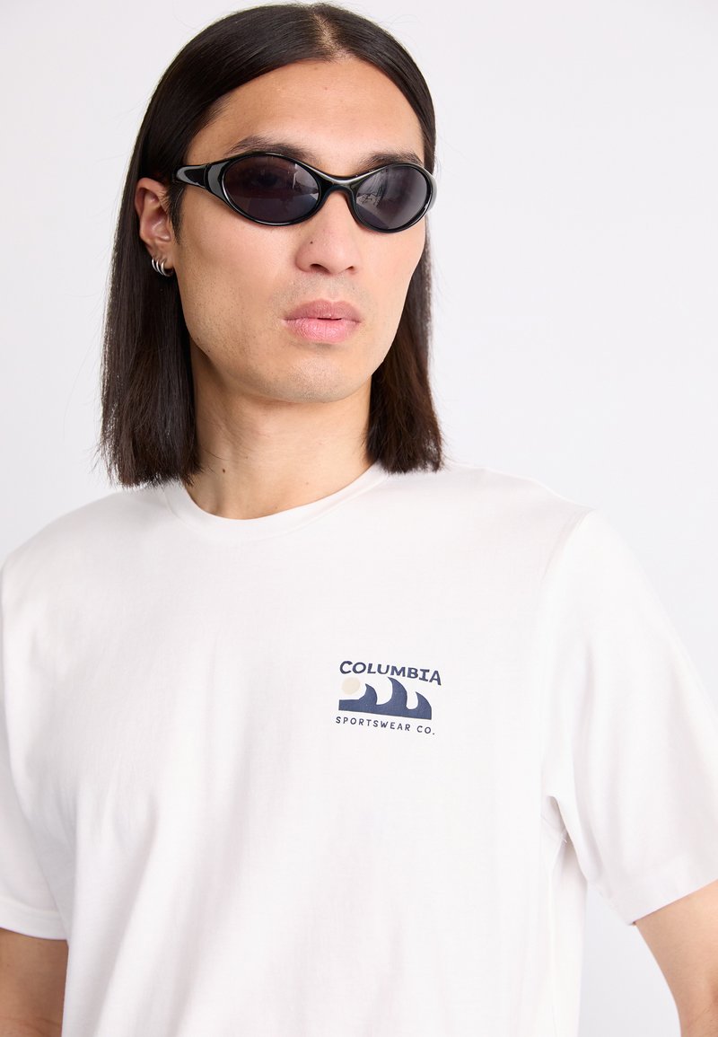 Czarne owalne okulary przeciwsłoneczne o smukłej ramce, połączone z białą koszulą typu crew-neck z granatowym logo i wzorem fali na piersi.