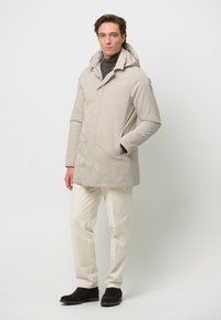 Beige vattentät jacka med huva, med rak skärning och två sidofickor. Kombinerad med krämfärgad byxor och mörk mocka ankelstövlar.