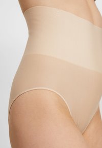 Beige høye livformende plagg med en glatt tekstur. Har en ribbet overdel og elastisk midjebånd for en sikker passform.