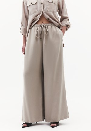 Beżowe szerokie spodnie z sznurkiem w pasie, w połączeniu z pasującą koszulą na guziki, noszone z czarnymi sandałami na obcasie z odkrytymi palcami. Ręce w kieszeniach.