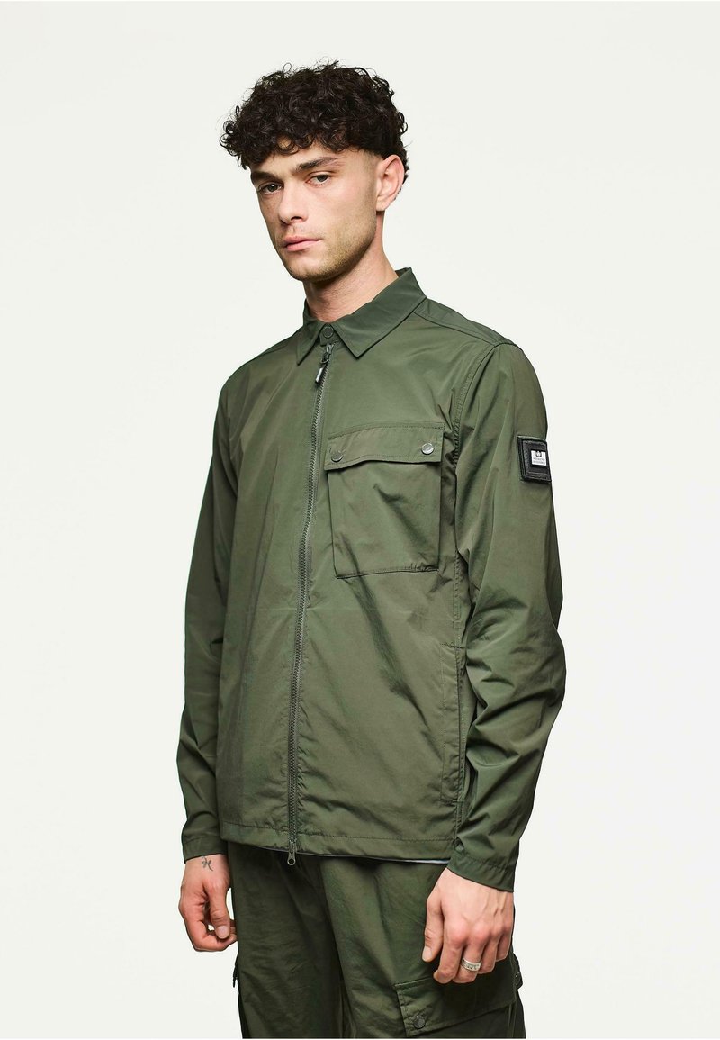 Chaqueta ligera verde con cuello, cremallera frontal y un bolsillo en el pecho. Presenta un parche de la marca en la manga y una textura suave.