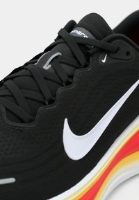 Chaussure de sport noire Nike avec logo swoosh blanc, lacets noirs et semelle multicolore dans des tons jaunes, rouges et blancs.