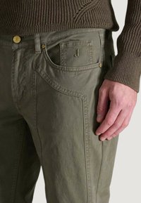 Los pantalones de algodón verde oliva presentan un acabado texturizado, bolsillos curvados y un sutil detalle del logo en el bolsillo delantero. Incluyen detalles de botones.