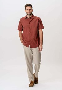 Mand går fremad iført en rustfarvet kortærmet skjorte med knapper, beige bukser og brune snørebåndssko, hænderne i lommerne.